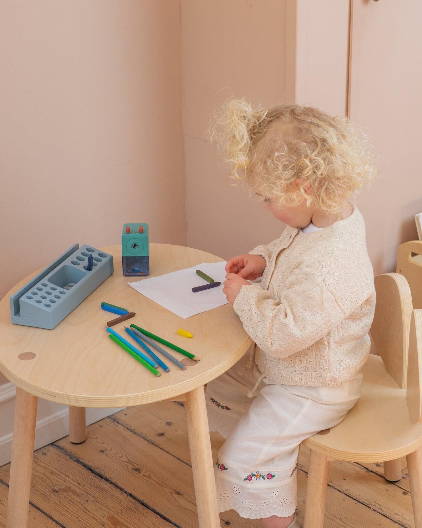 Child sitting at a small wooden table with art supplies, drawing on paper.