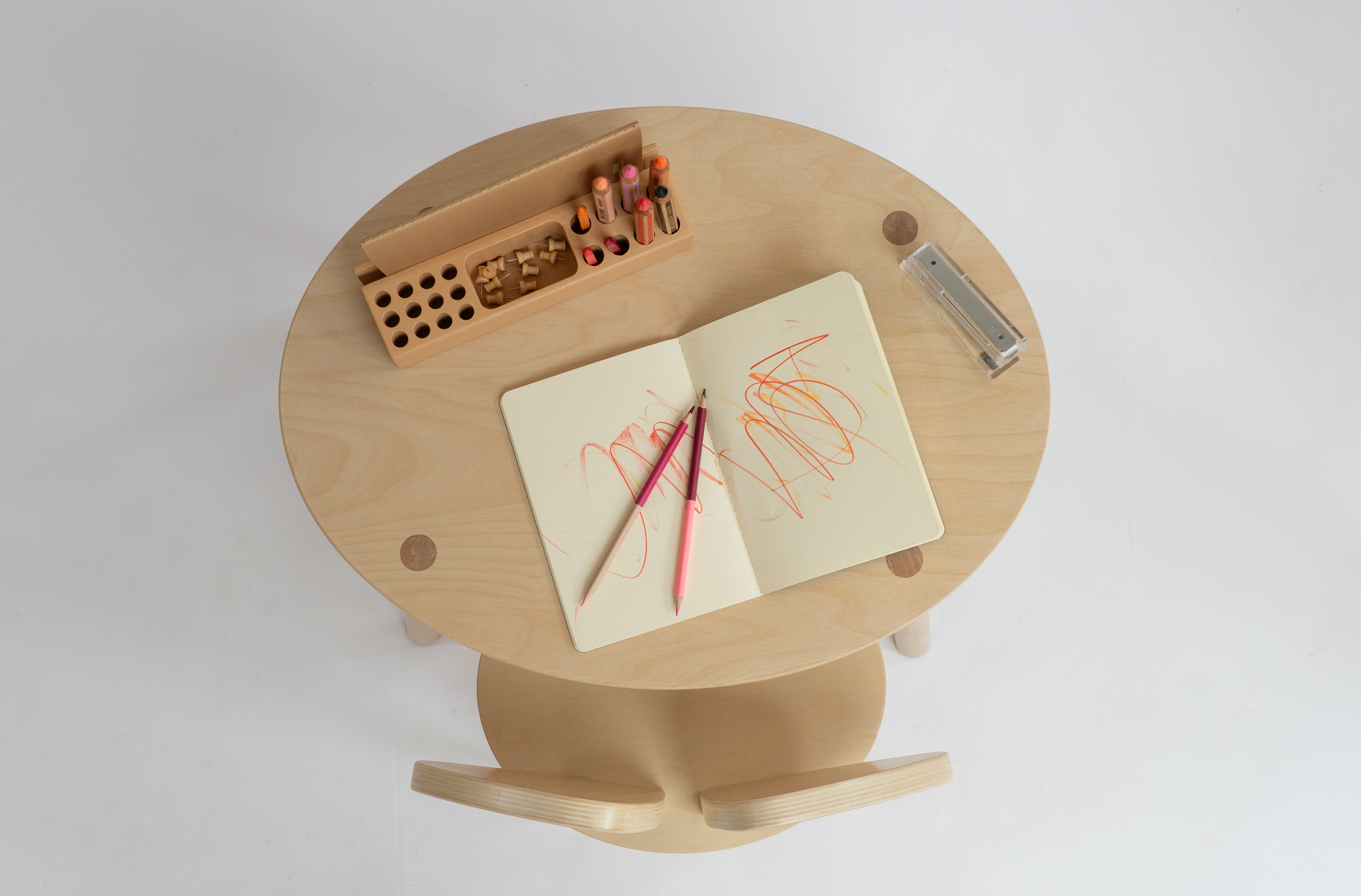A top-down view of a light natural wood children's desk with a matching curved chair. On the desk are an open notebook with orange scribbles, two pink and purple pencils, a wooden pencil holder with colored pencils, and a small clear plastic case, all set against a clean white background.