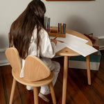 Person sitting at a wooden table with a chair, surrounded by art supplies in a room with a painting on the wall.