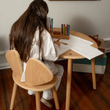 Person sitting at a wooden table with a chair, surrounded by art supplies in a room with a painting on the wall.