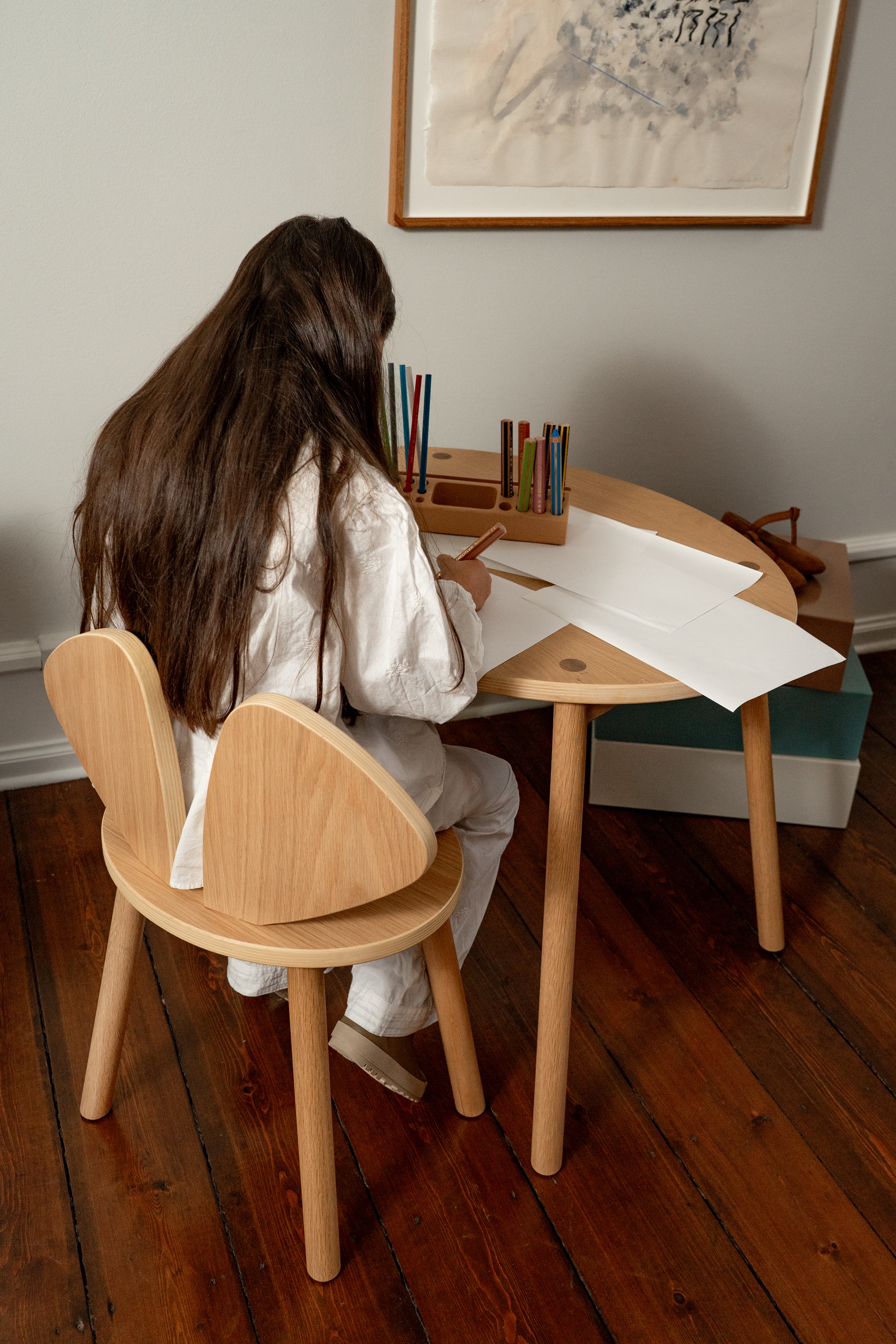 Person sitting at a wooden table with a chair, surrounded by art supplies in a room with a painting on the wall.