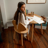 Young girl sitting at a small wooden table with art supplies, drawing on paper.