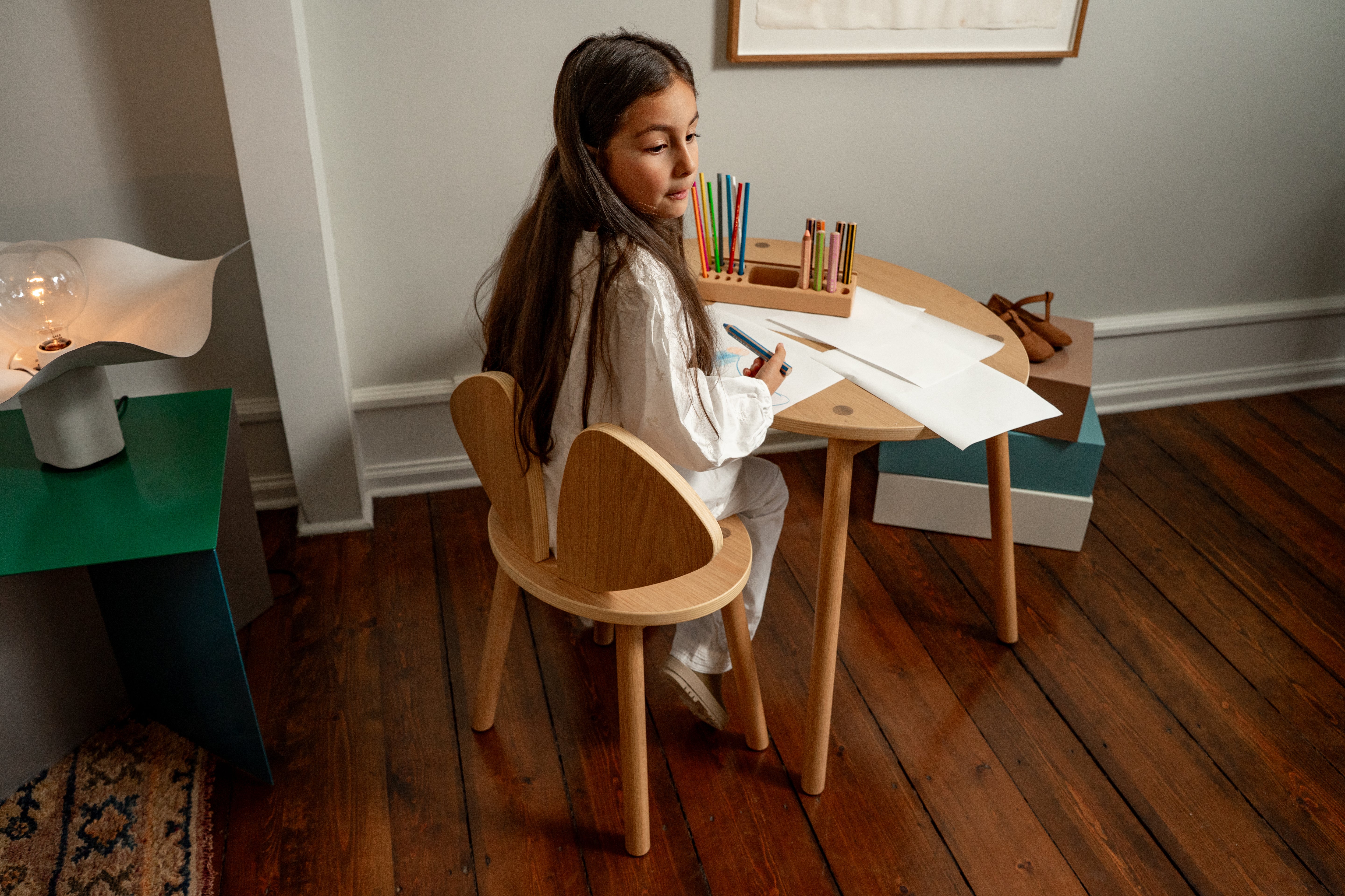 Young girl sitting at a small wooden table with art supplies, drawing on paper.