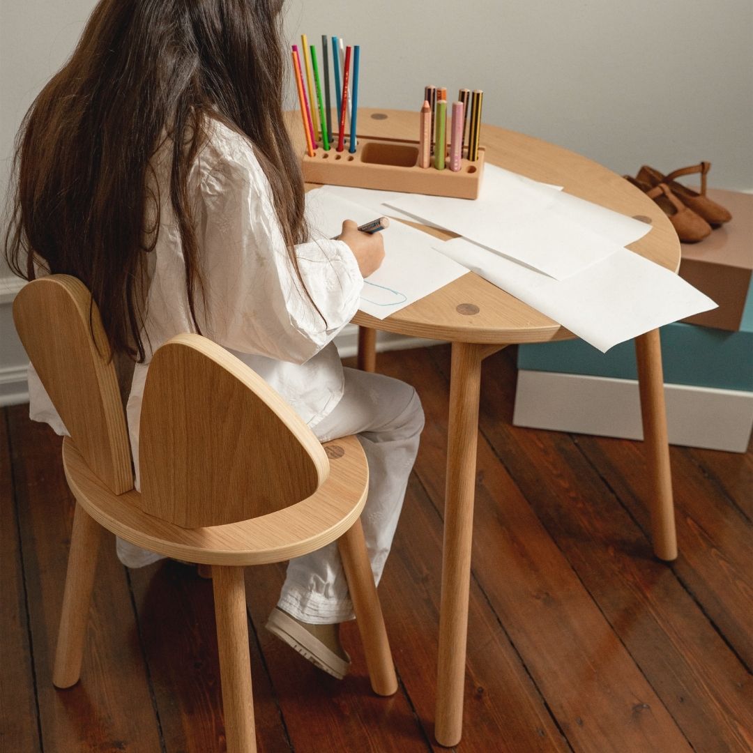 Child sitting at a small wooden table with papers and colored pencils, wearing a white shirt and light-colored pants.