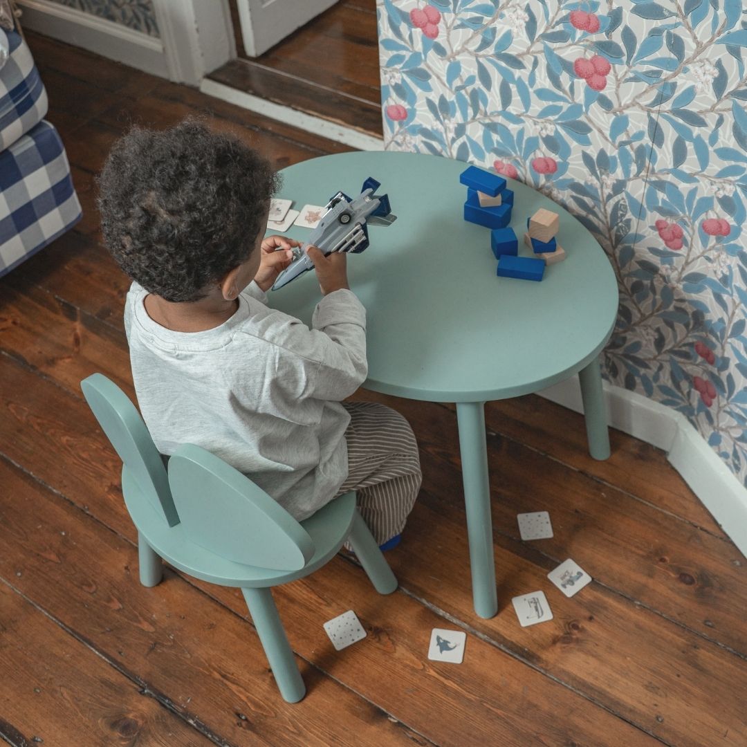 Child playing with toys at a green table in a room with wooden flooring and floral wallpaper.