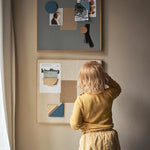 Child in a yellow shirt and shorts standing in front of a wall with framed pictures and a bulletin board.