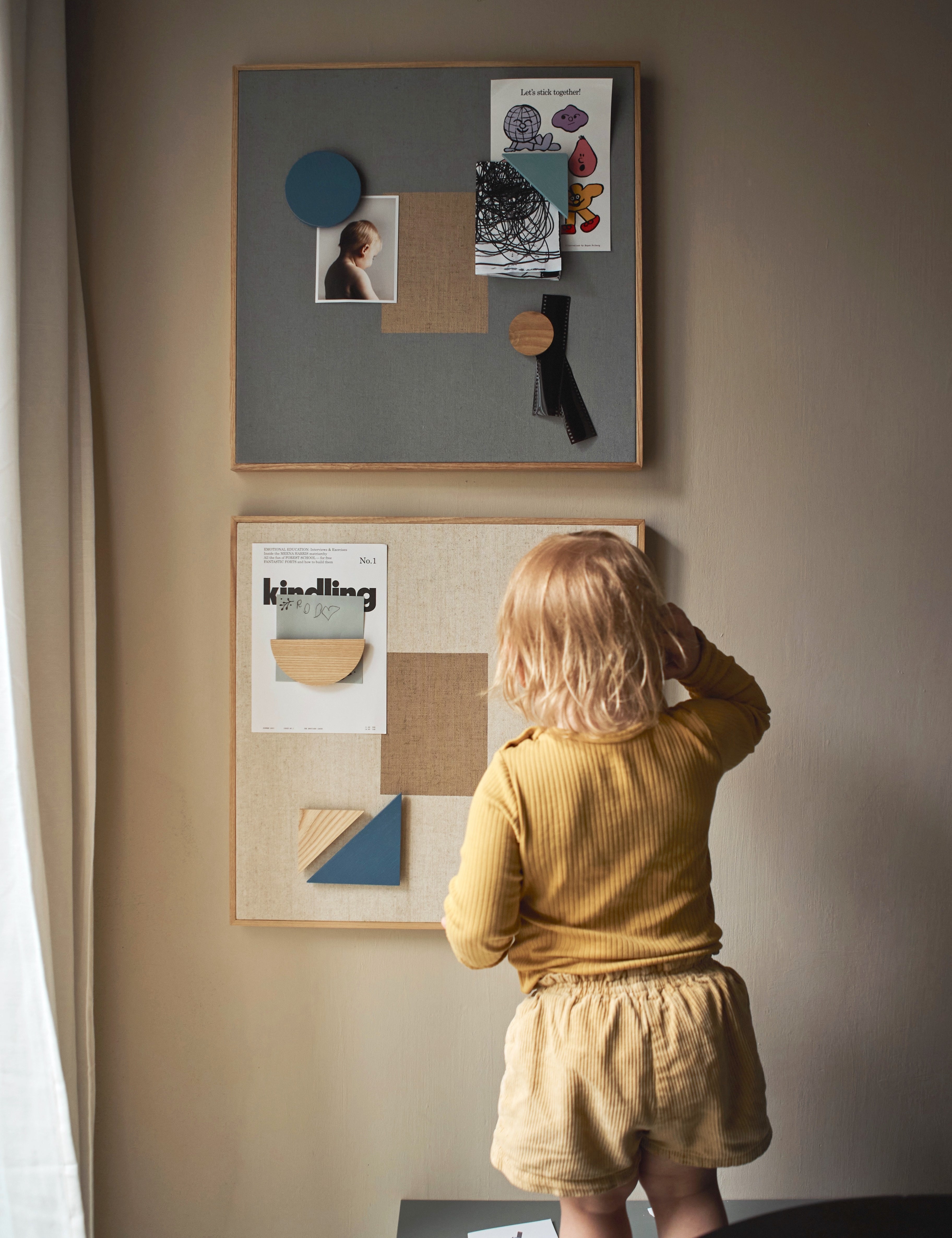 Child in a yellow shirt and shorts standing in front of a wall with framed pictures and a bulletin board.