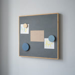 Gray bulletin board with wooden frame on a gray wall, featuring various notes and magnets.
