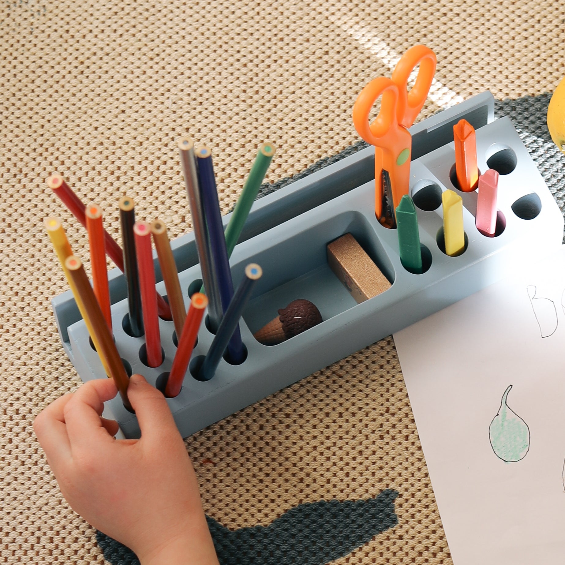 Children's art supplies including colored pencils, scissors, and crayons in a holder on a textured surface.