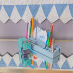 A light blue wooden pencil holder and sharpener organizer mounted on a wooden shelf, holding various colored pencils and paper. The background features a patterned quilt with blue and white triangles.