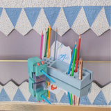 A light blue wooden pencil holder and sharpener organizer mounted on a wooden shelf, holding various colored pencils and paper. The background features a patterned quilt with blue and white triangles.
