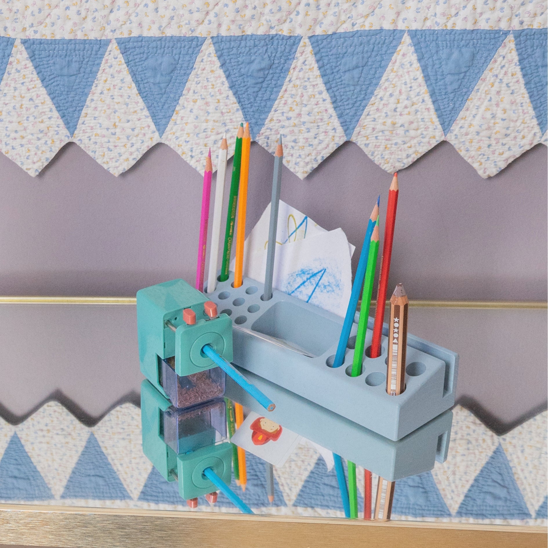 A light blue wooden pencil holder and sharpener organizer mounted on a wooden shelf, holding various colored pencils and paper. The background features a patterned quilt with blue and white triangles.