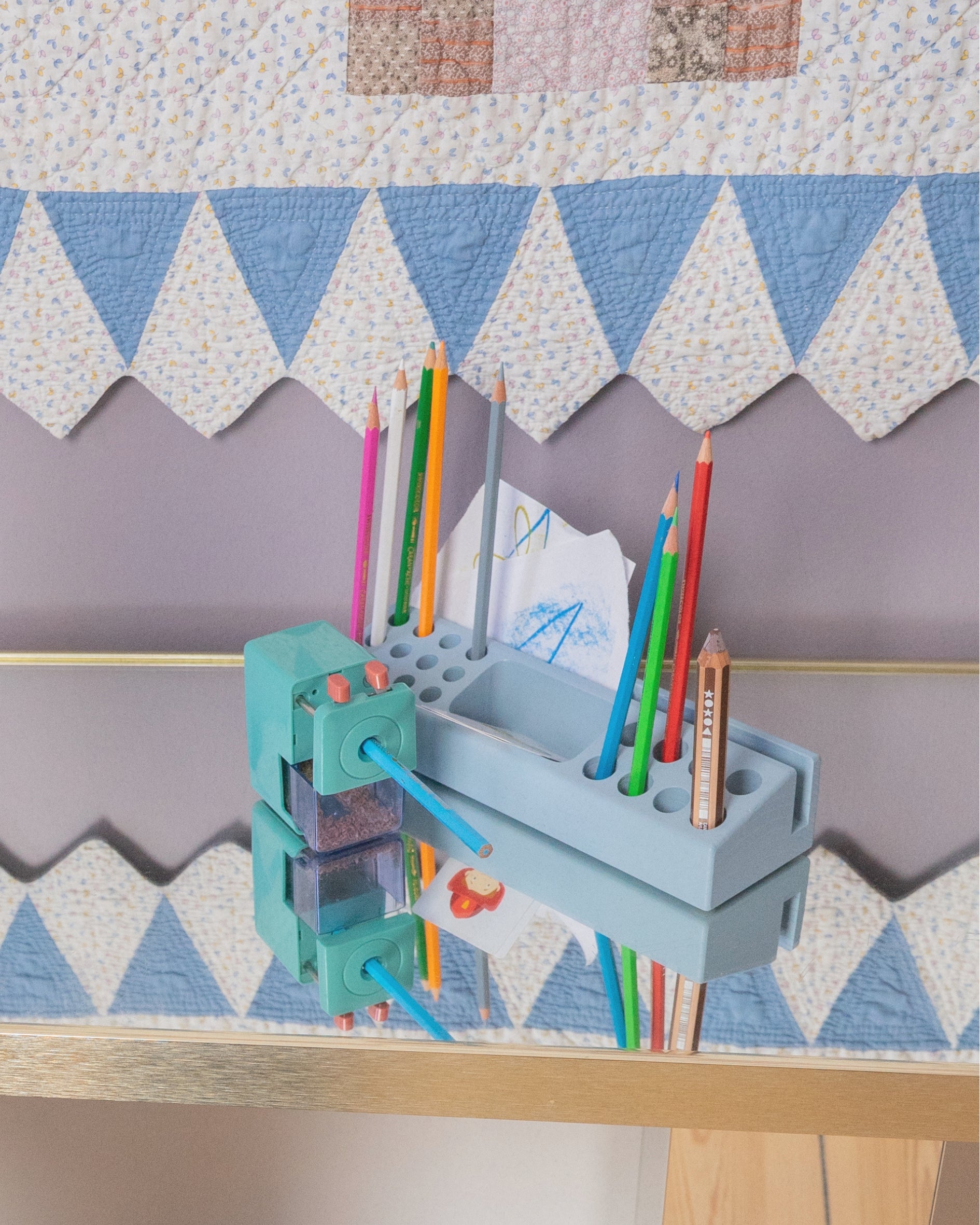 A light blue wooden pencil holder and sharpener organizer mounted on a wooden shelf, holding various colored pencils and paper. The background features a patterned quilt with blue and white triangles.