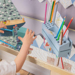 A child's hand reaches toward a light blue wooden toy desk organizer holding colorful pencils, with a matching toy boat and patterned quilted wall in the background.