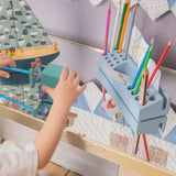 A child's hand reaches toward a light blue wooden toy desk organizer holding colorful pencils, with a matching toy boat and patterned quilted wall in the background.