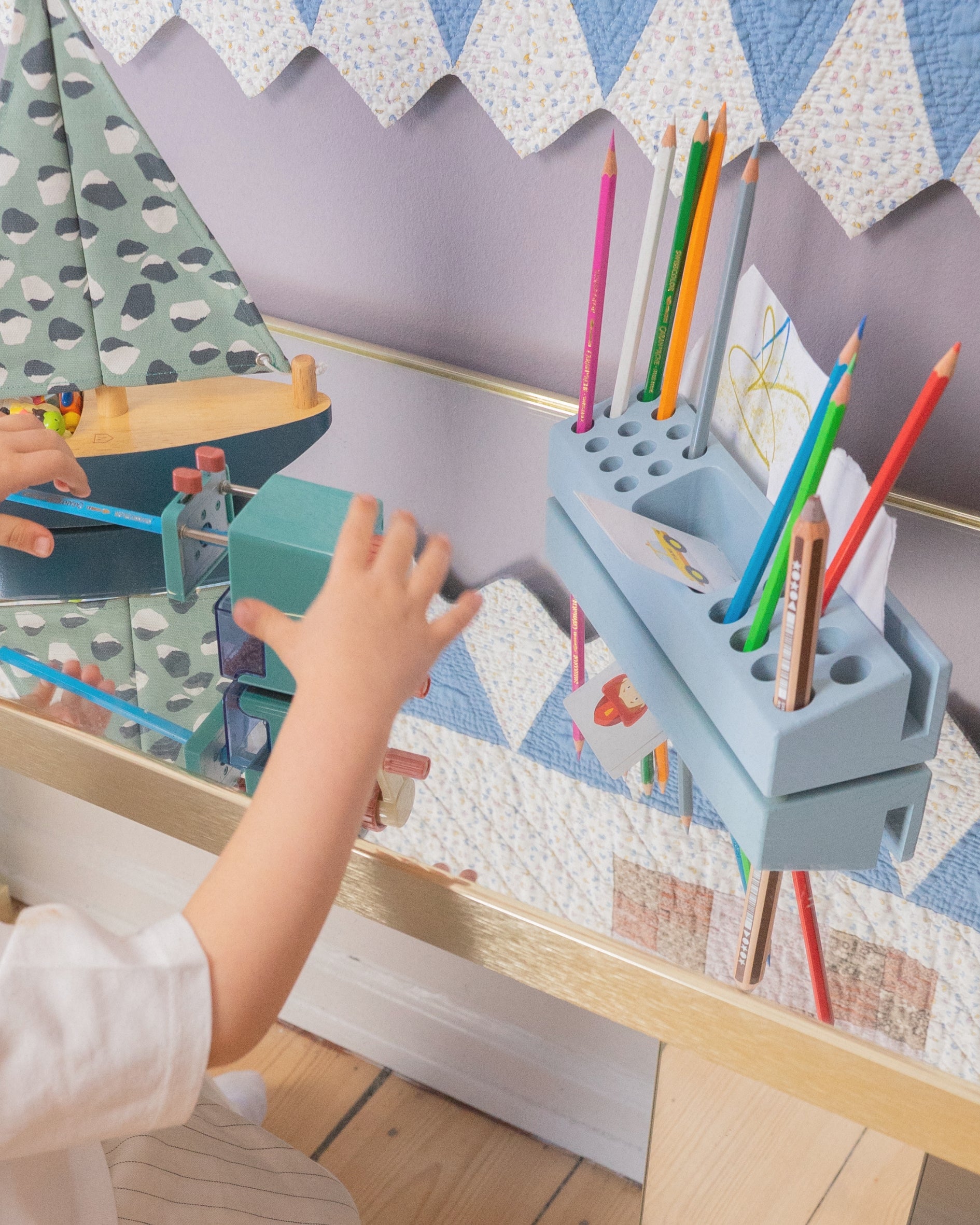 A child's hand reaches toward a light blue wooden toy desk organizer holding colorful pencils, with a matching toy boat and patterned quilted wall in the background.