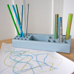 Blue pencil holder with colorful pens and a child's drawing on a white sheet of paper.