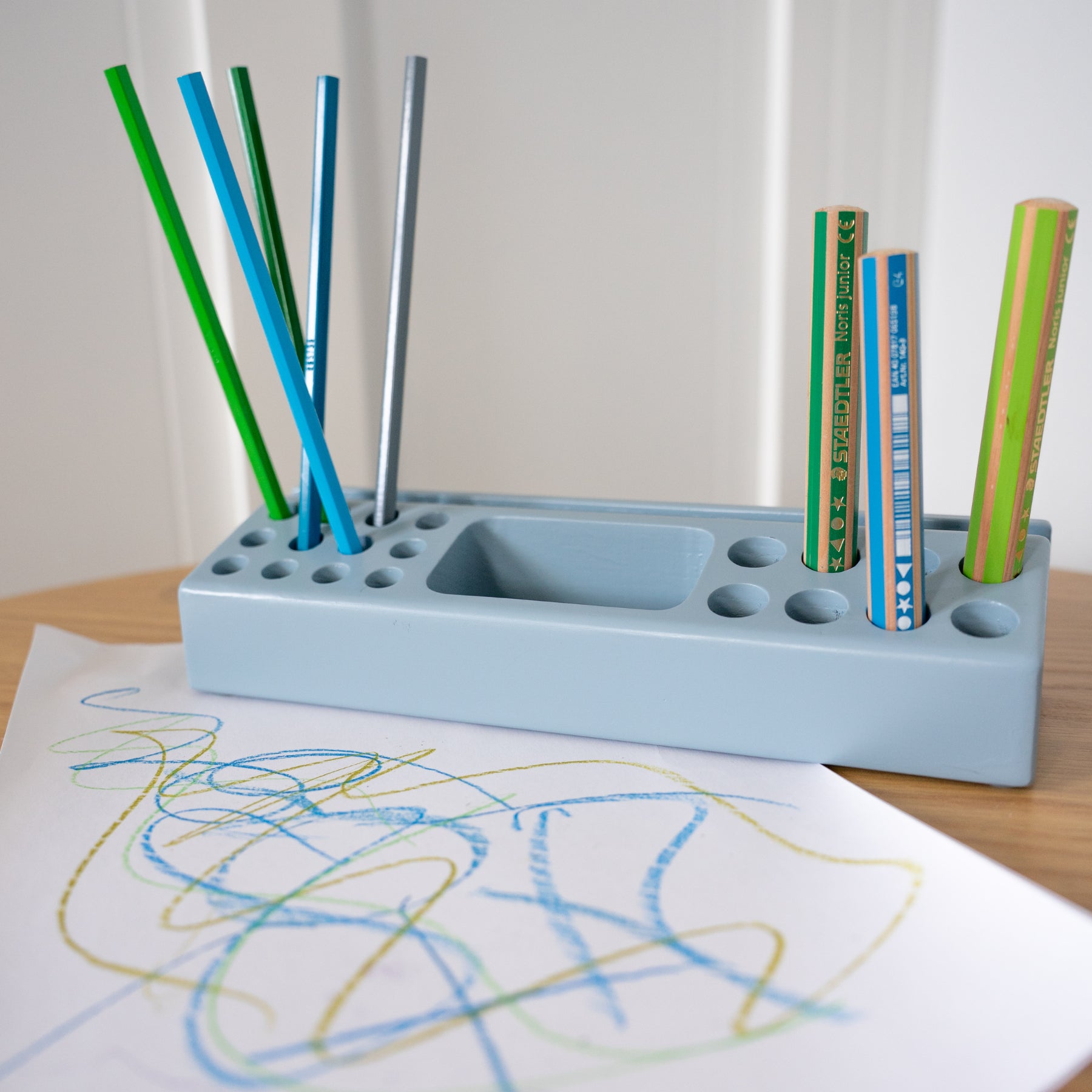 Blue pencil holder with colorful pens and a child's drawing on a white sheet of paper.