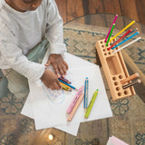 Child drawing with colored pencils on a piece of paper, with a wooden pencil holder filled with colorful pencils on a glass table.