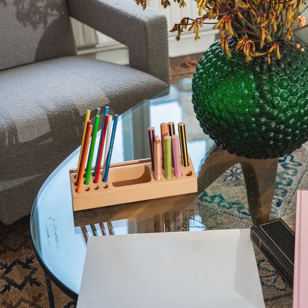 Colorful pens in a holder on a glass table with a green textured vase and plant in the background.