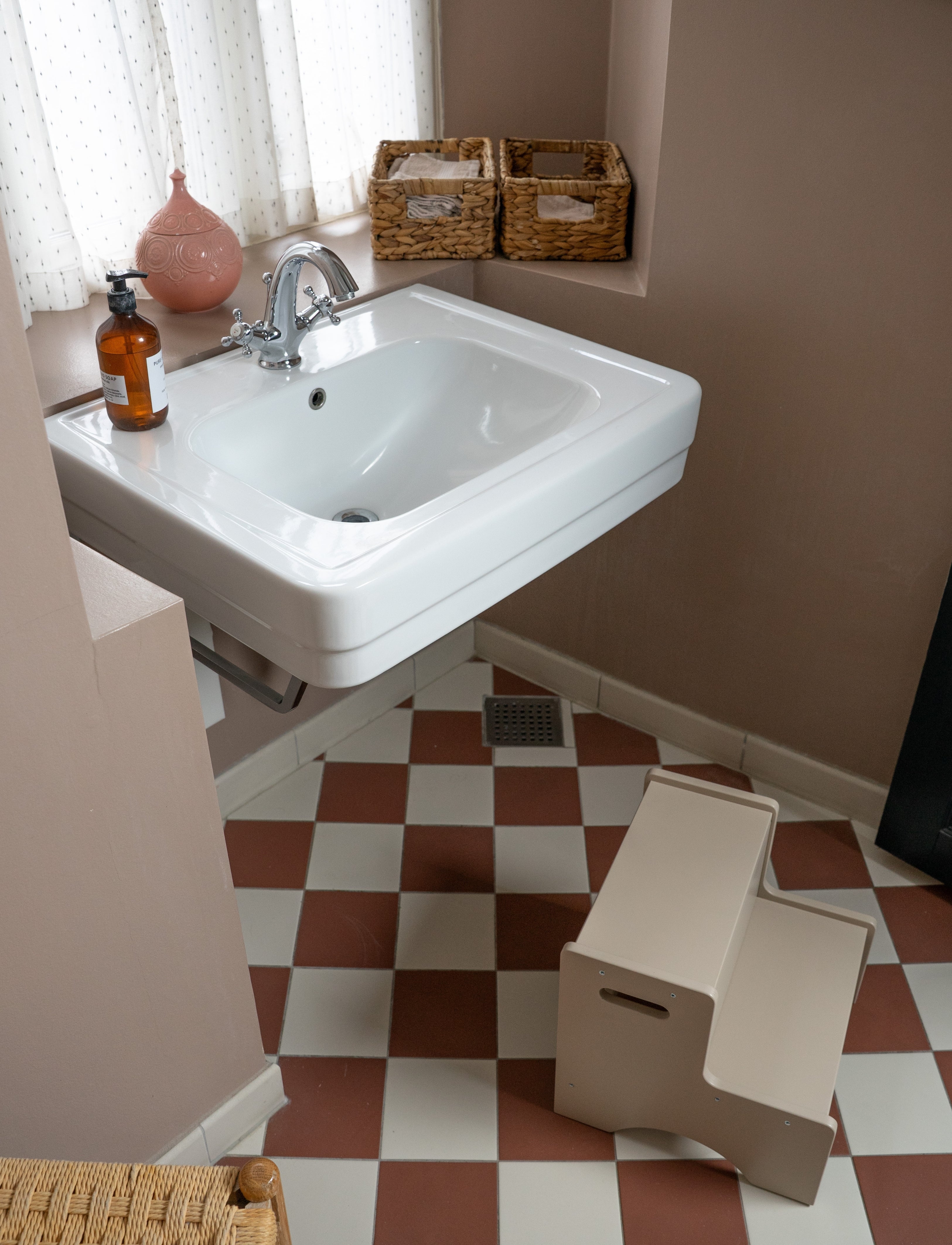A white wall-mounted sink with a chrome faucet in a bathroom, shown with a beige step stool, checkered floor, and wicker baskets on the windowsill.