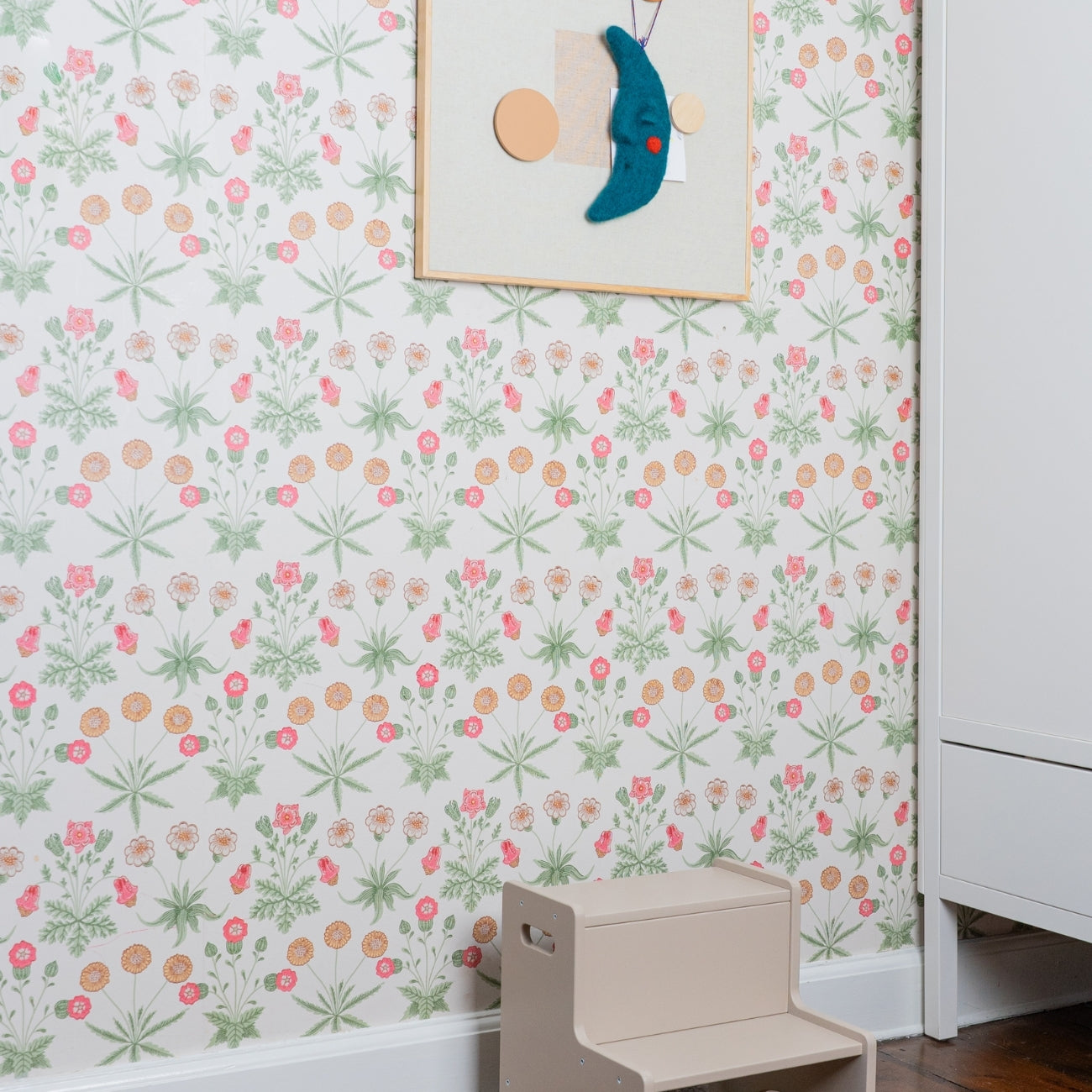 Children's step stool against a floral wallpapered wall with a framed picture.
