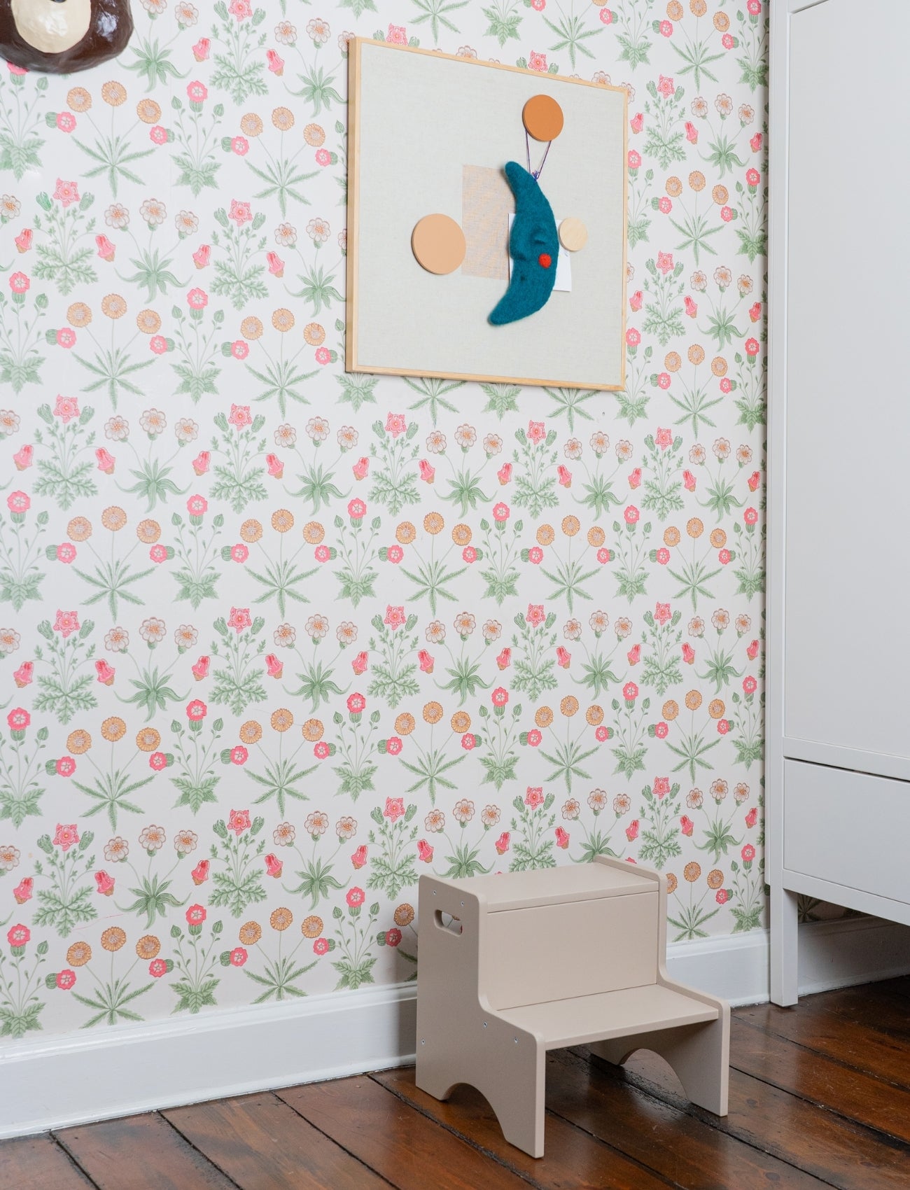 Children's step stool against a floral wallpapered wall with a framed picture.