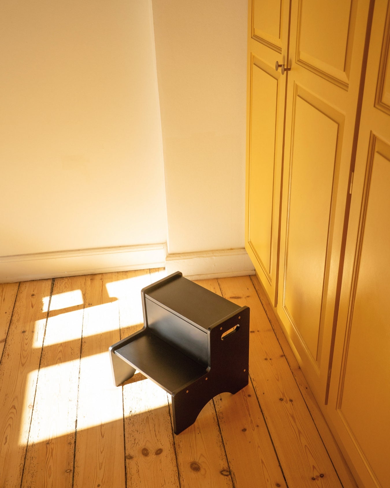 Small black step stool on a wooden floor next to a white wall and yellow cabinet.