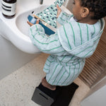 Child in a green and white striped bathrobe using a step stool to reach a sink.