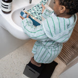Child in a green and white striped bathrobe using a step stool to reach a sink.