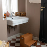 A brown wooden two-step stool placed on a brown and white checkered floor in a bathroom setting with a white sink, brown walls, and a black door.