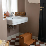 A brown wooden two-step stool placed on a brown and white checkered floor in a bathroom setting with a white sink, brown walls, and a black door.