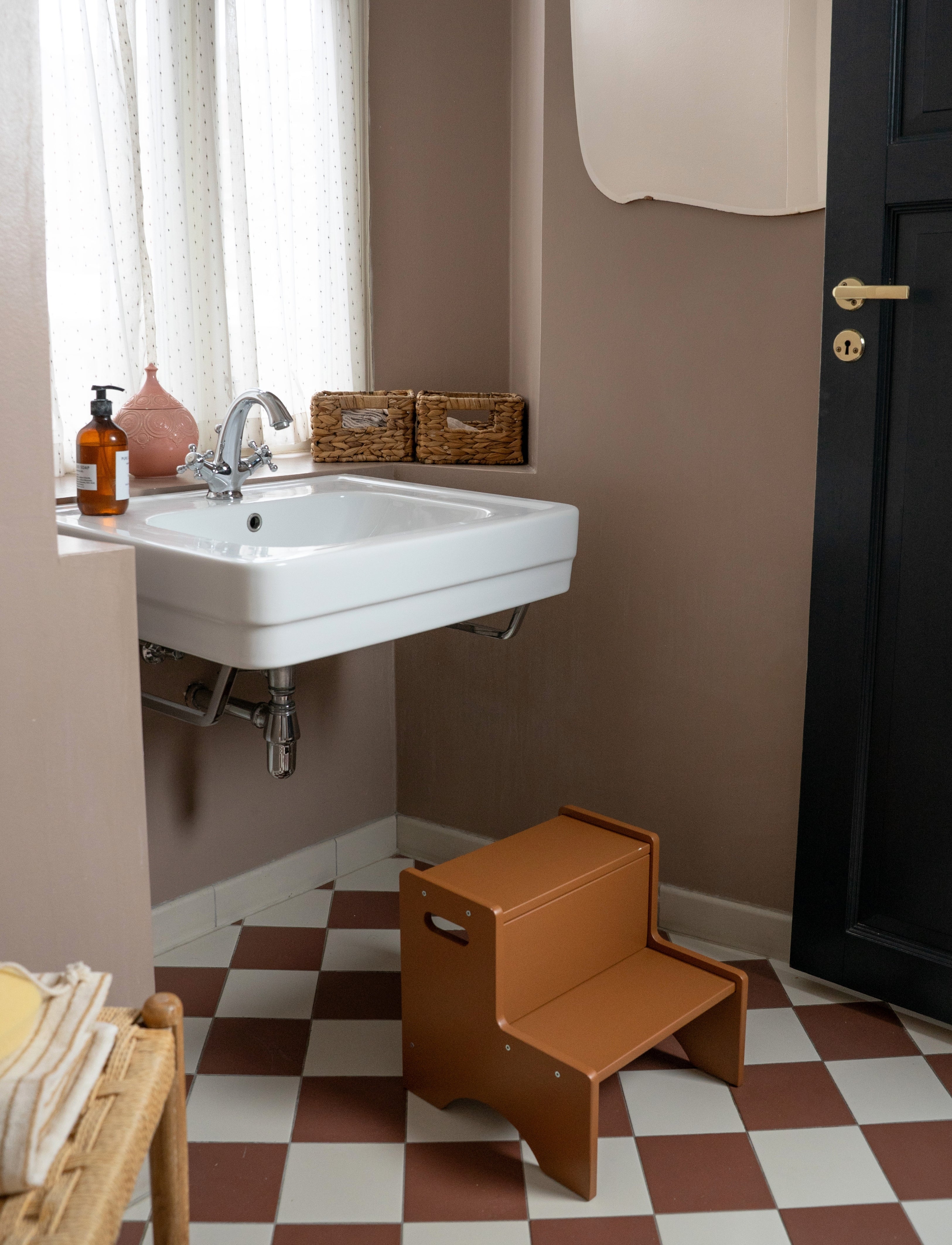 A brown wooden two-step stool placed on a brown and white checkered floor in a bathroom setting with a white sink, brown walls, and a black door.