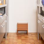 Small wooden stool in a kitchen with hexagonal wooden floor tiles and white walls.