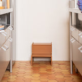 Small wooden stool in a kitchen with hexagonal wooden floor tiles and white walls.