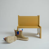 A pair of beige Chelsea-style rain boots with blue and orange trim, displayed next to a minimalist mustard yellow bench on a light gray studio background.