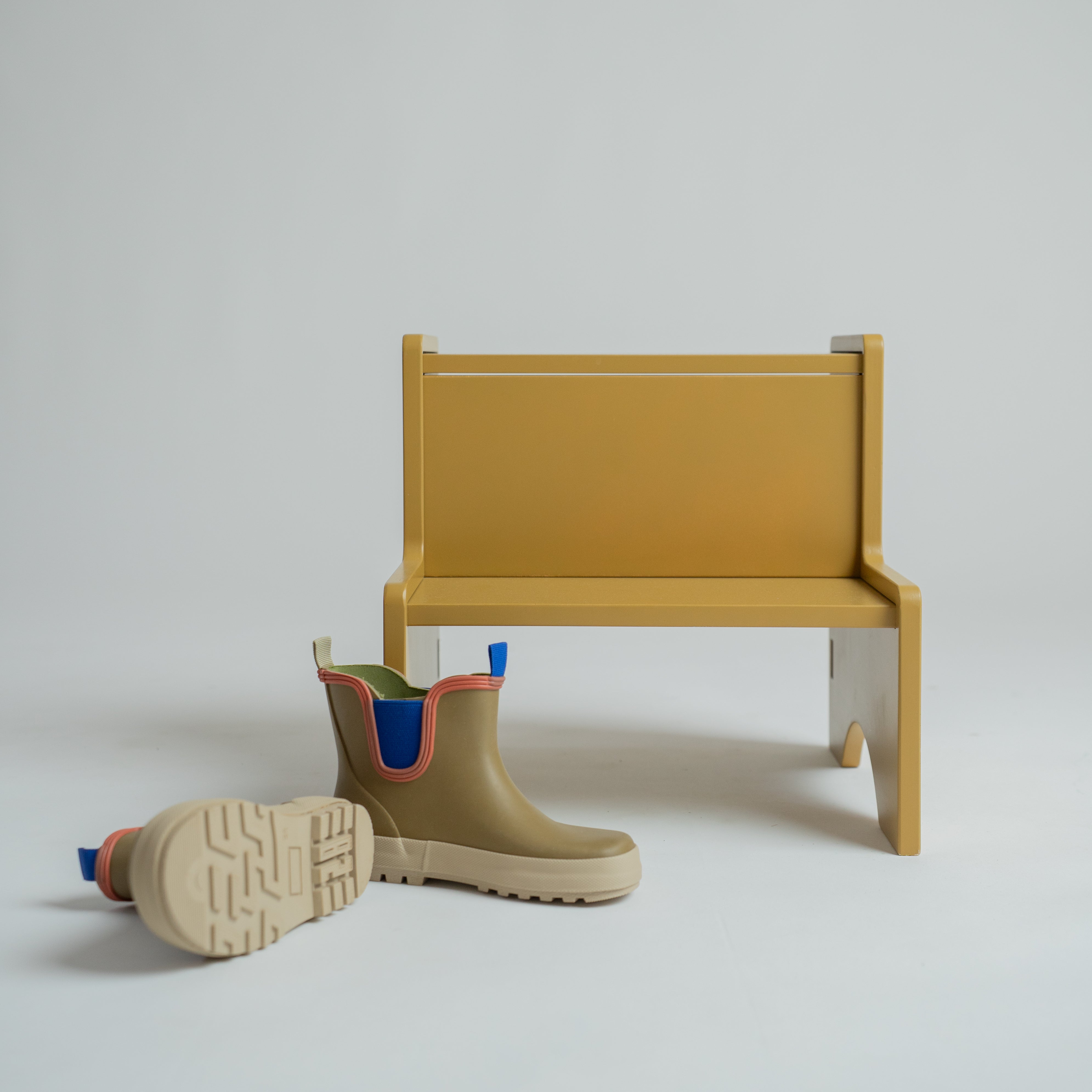 A pair of beige Chelsea-style rain boots with blue and orange trim, displayed next to a minimalist mustard yellow bench on a light gray studio background.
