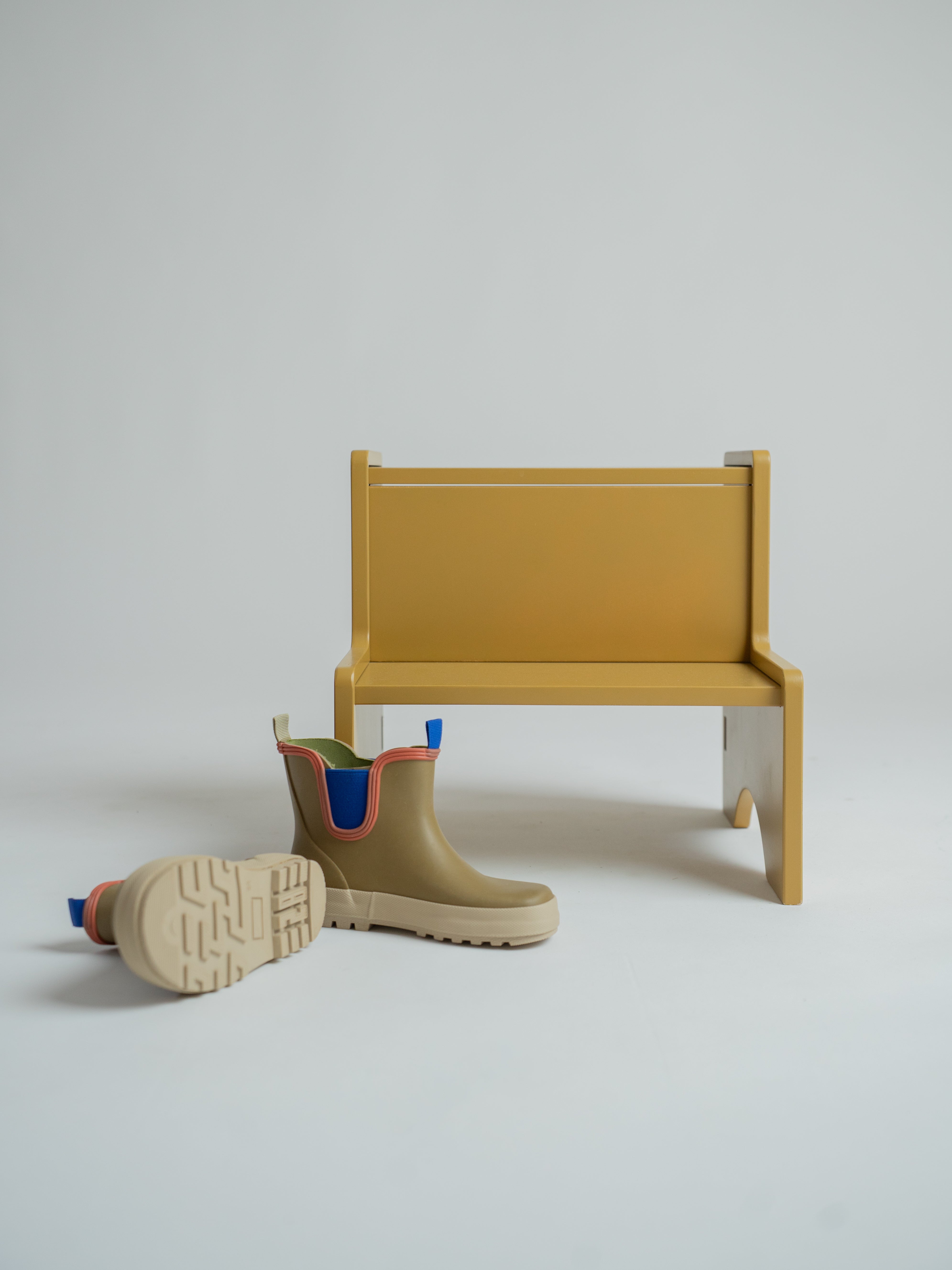 A pair of beige Chelsea-style rain boots with blue and orange trim, displayed next to a minimalist mustard yellow bench on a light gray studio background.