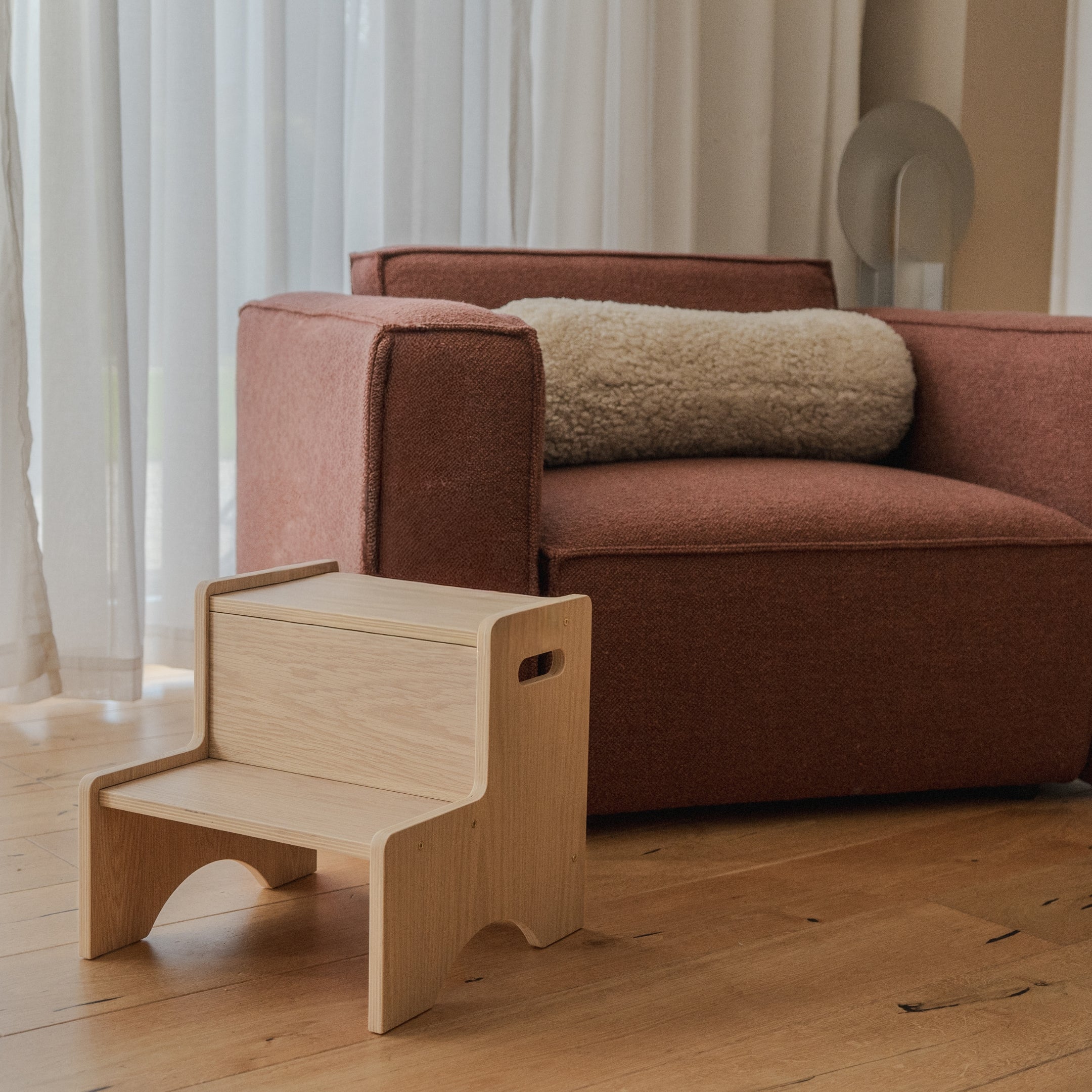 Wooden step stool next to a brown sofa with a beige cushion in a room with curtains.