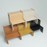 Four minimalist wooden step stools in light natural wood, warm brown, mustard yellow, and matte black, arranged in a stacked, overlapping display on a light gray background.