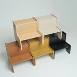 Four minimalist wooden step stools in light natural wood, warm brown, mustard yellow, and matte black, arranged in a stacked, overlapping display on a light gray background.