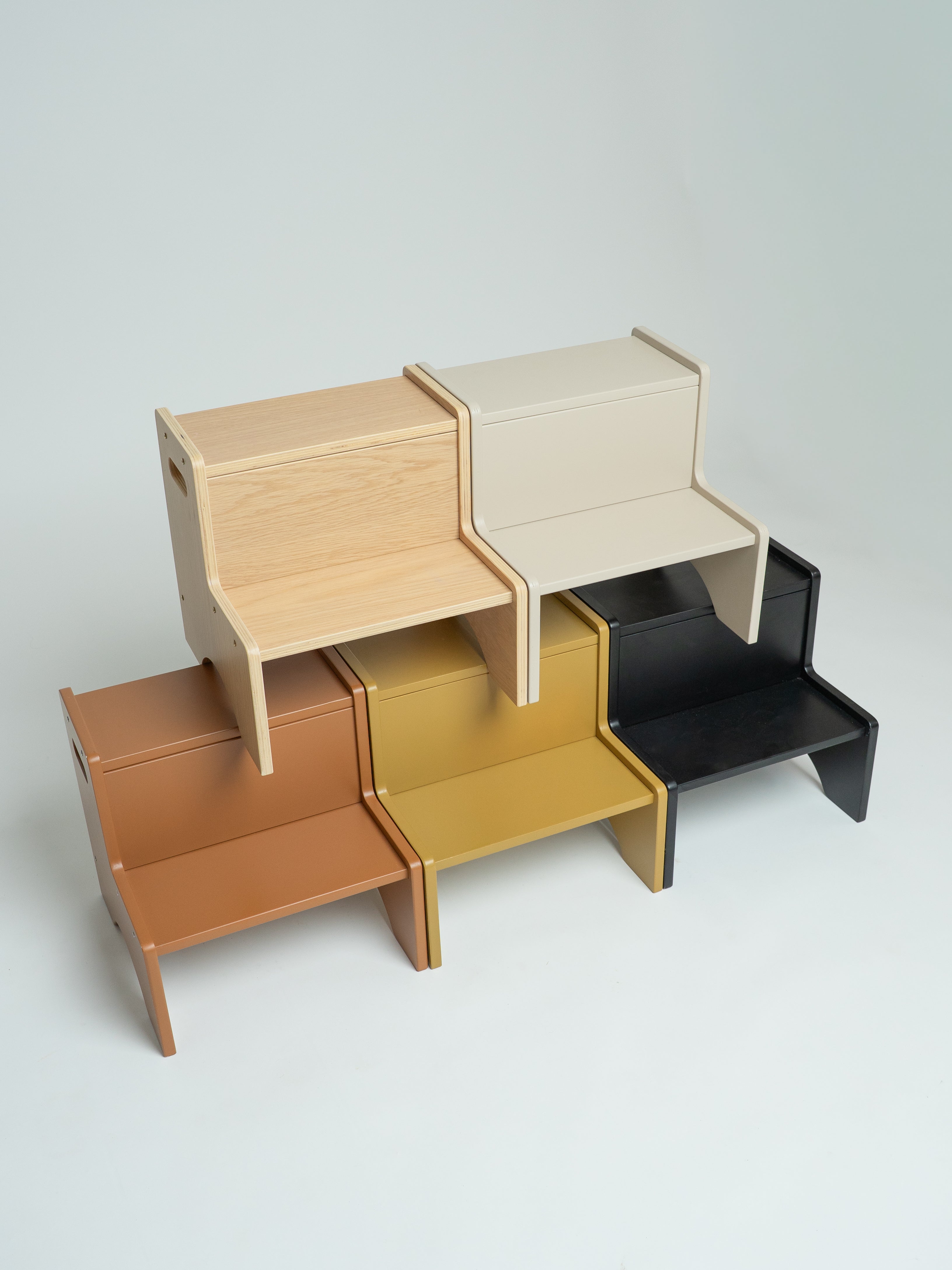 Four minimalist wooden step stools in light natural wood, warm brown, mustard yellow, and matte black, arranged in a stacked, overlapping display on a light gray background.