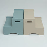 Two minimalist two-step platforms shown side by side on a light gray studio background. The left step is a muted dusty blue, and the right step is a warm beige.