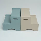 Two minimalist two-step platforms shown side by side on a light gray studio background. The left step is a muted dusty blue, and the right step is a warm beige.