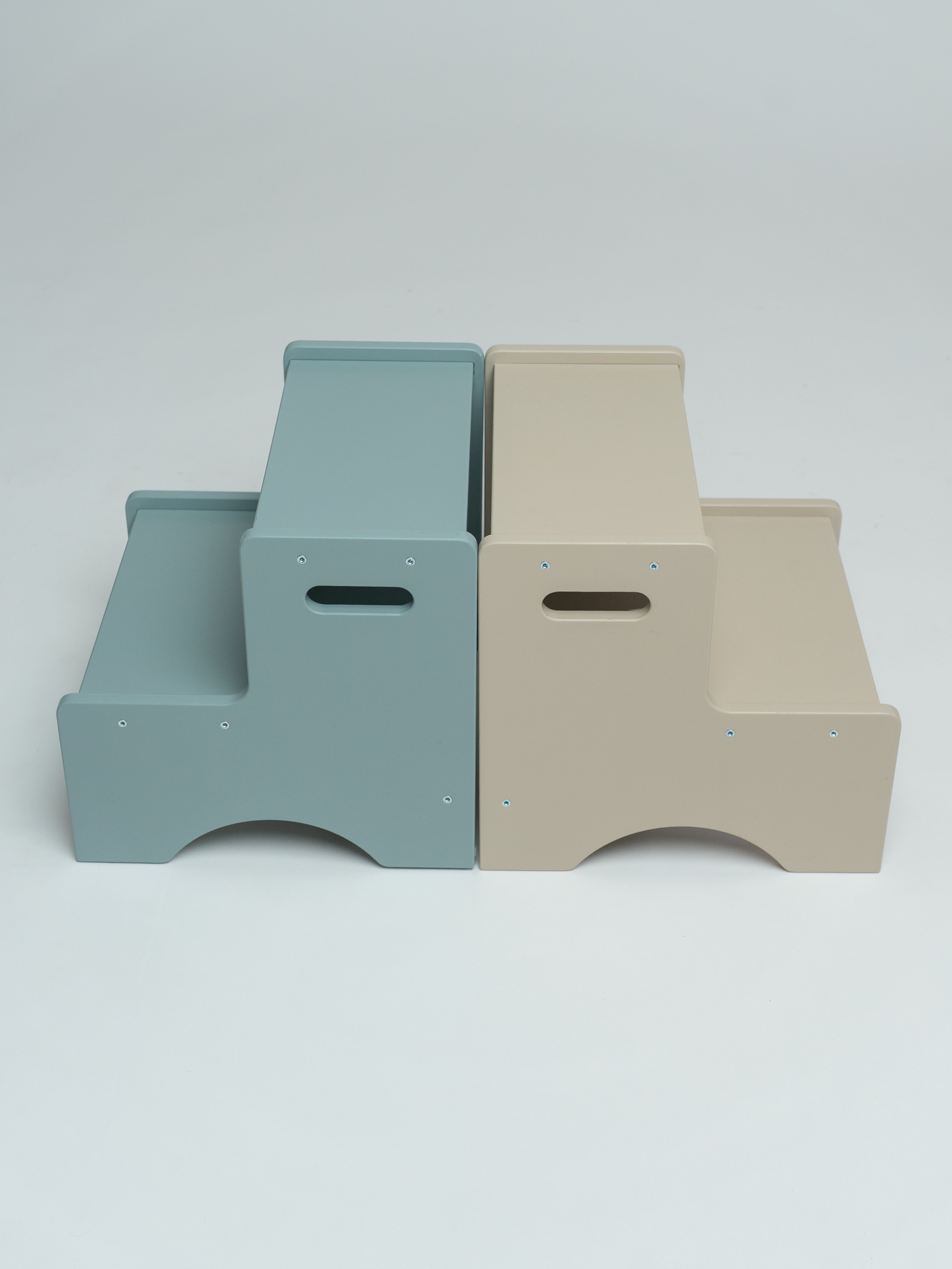 Two minimalist two-step platforms shown side by side on a light gray studio background. The left step is a muted dusty blue, and the right step is a warm beige.
