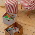 Two storage boxes on a wooden floor with striped ottoman and chair in the background.