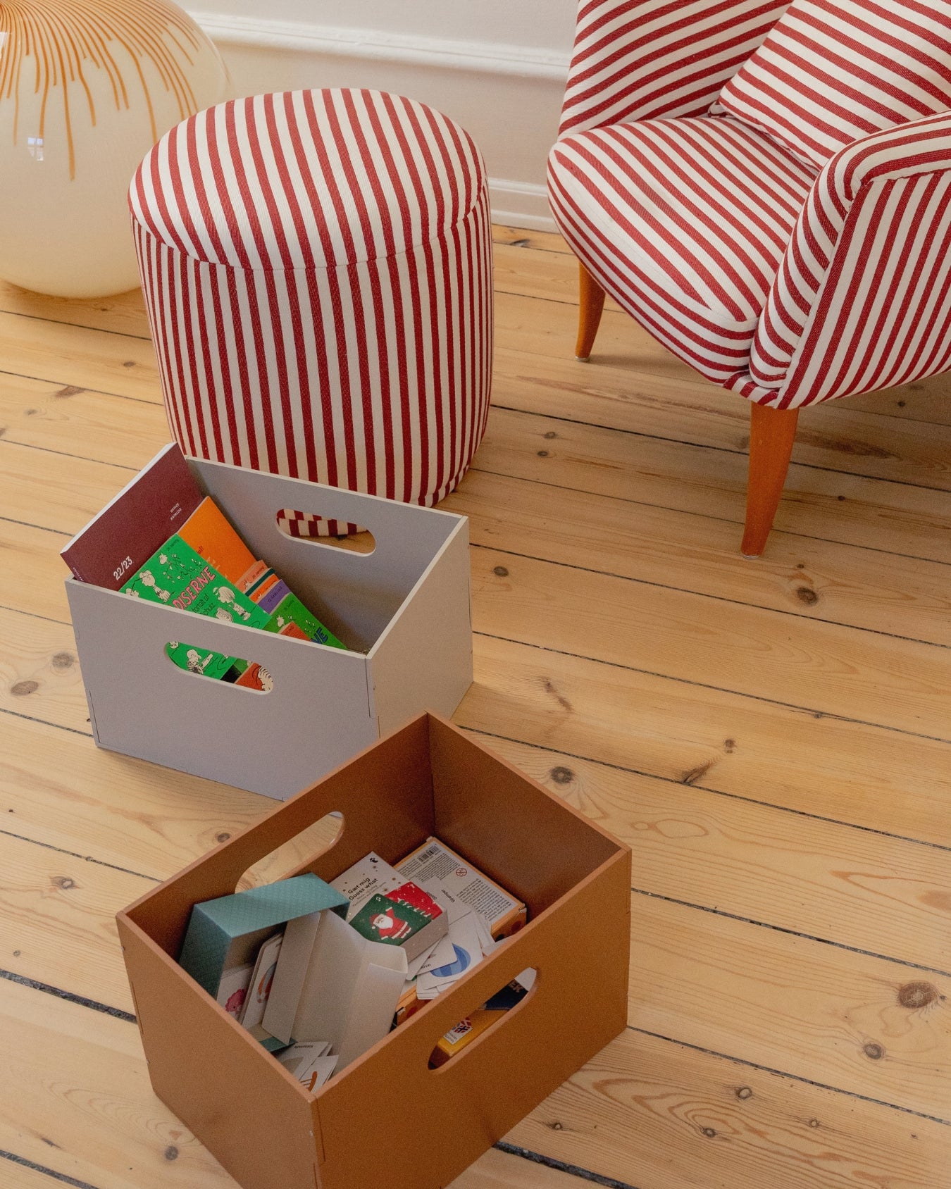Two storage boxes on a wooden floor with striped ottoman and chair in the background.