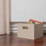 Beige storage box with colorful items on a wooden floor, curtains in the background