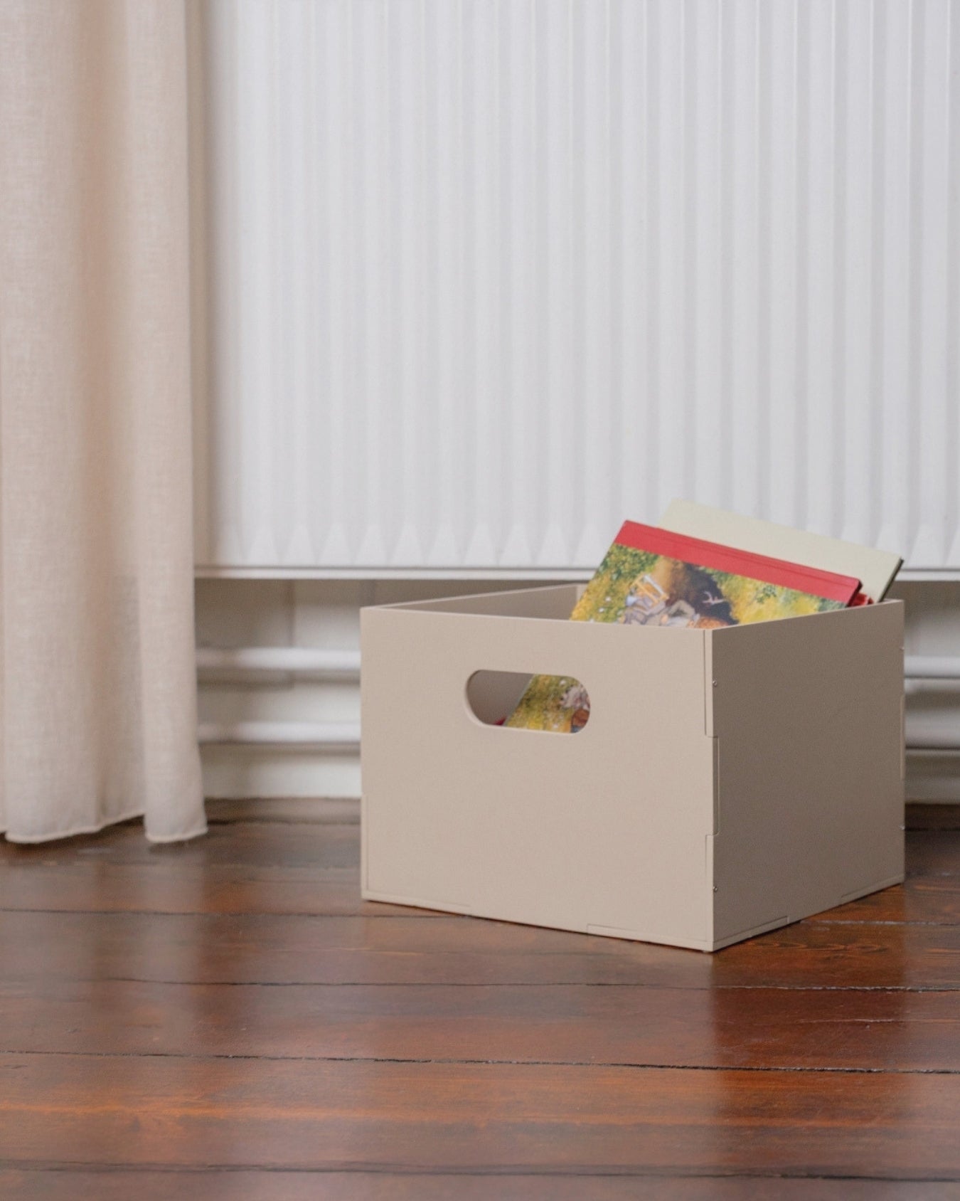 Beige storage box with colorful items on a wooden floor, curtains in the background