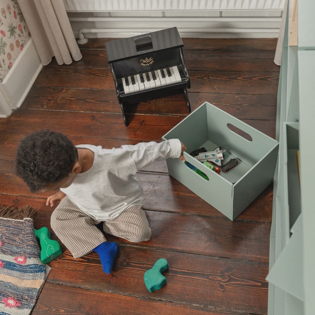 Child playing with a toy piano and a box of toys on a wooden floor.